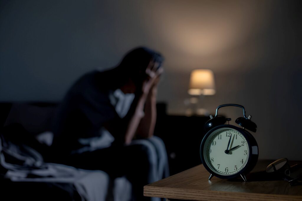 A person sitting on a bed with hands on head, looking stressed, next to an alarm clock showing 2:00, with a dim lamp in the background, highlighting bad sleep issues.