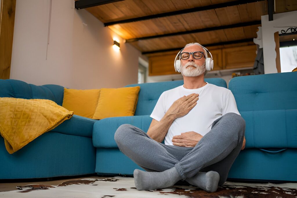 An elderly man practicing mindful habits, sitting cross-legged on the floor with headphones, hands on chest, in a cozy living room with a blue sofa and yellow cushions.